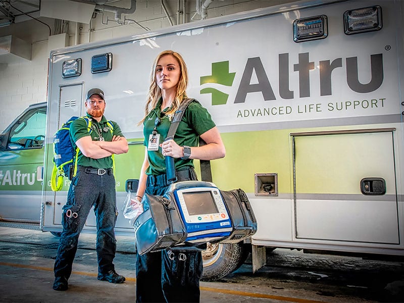 A professional photo of two Altru Hospital emergency medical technicians standing in front of an ambulance