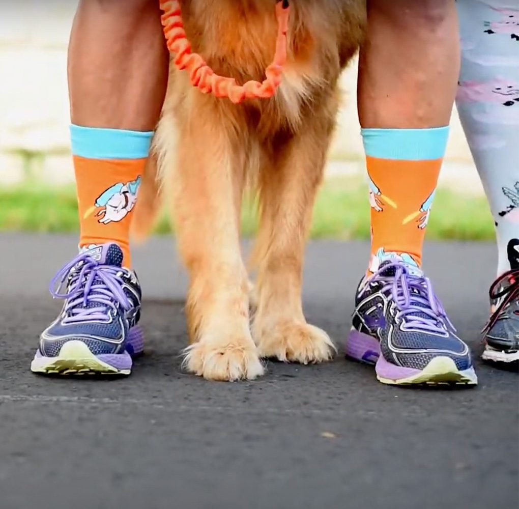Persons legs shot from knees down with orange socks and purple tennis shoes. A Golden Retriever stands between their legs.