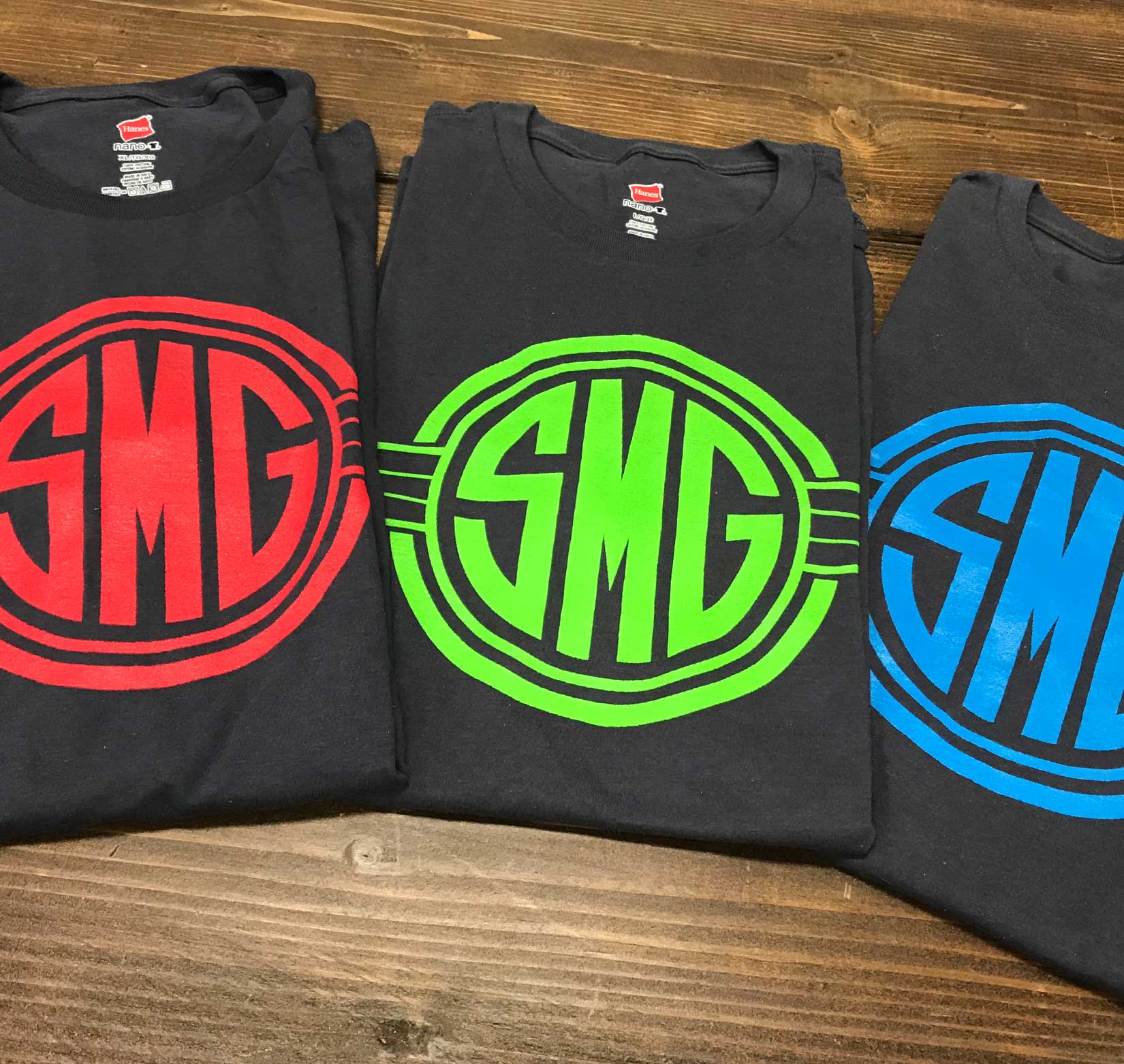 Three folded gray t-shirts with different colors of Stray Media Group logos lying on a table