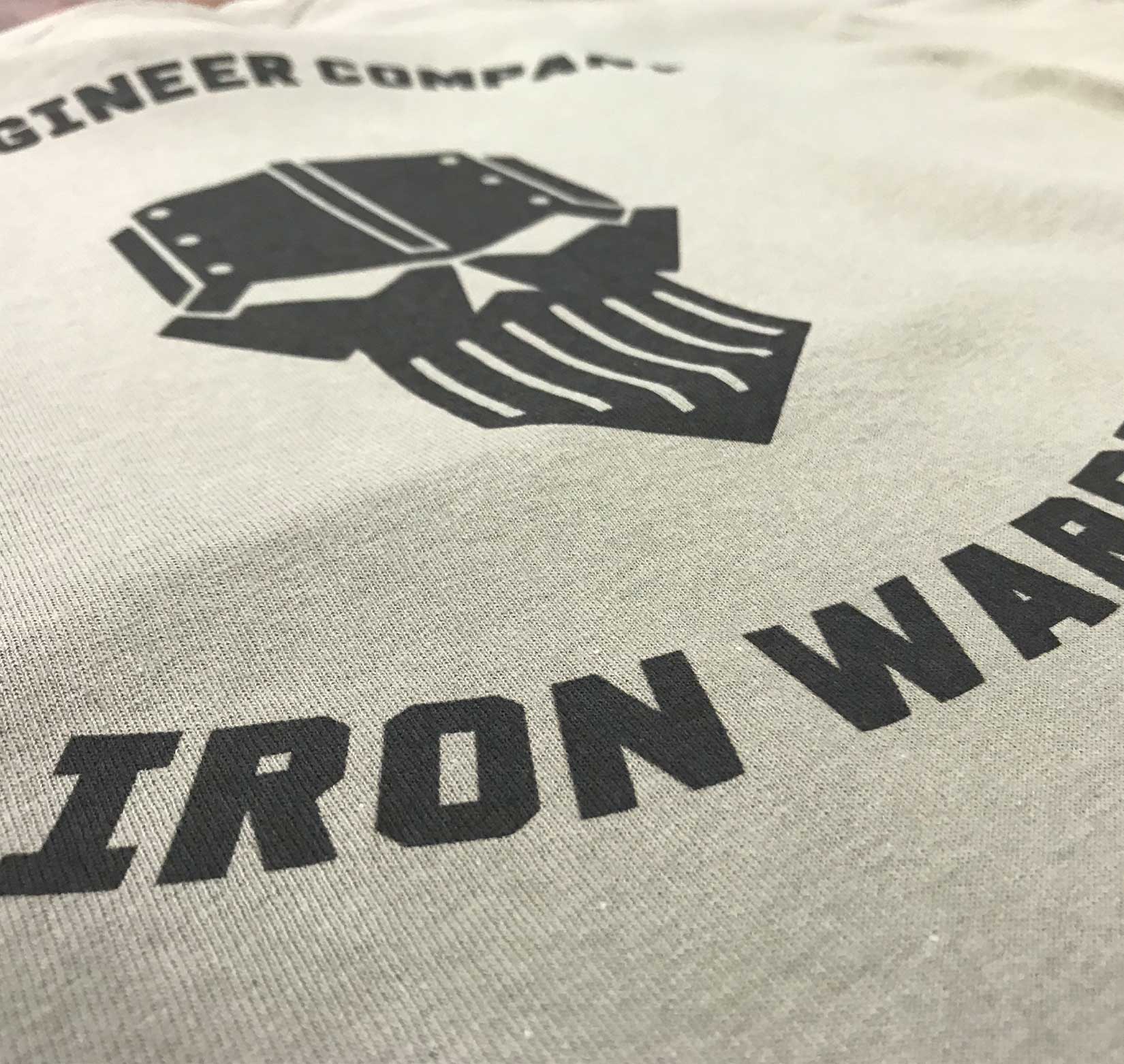 A closeup of a light tan t-shirt screen printed with a black logo that says “iron warriors” in all-caps text